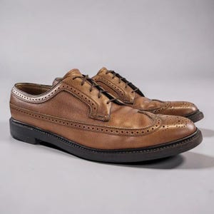May include: A pair of brown leather dress shoes with decorative perforations and black soles. The shoes have a classic wingtip design and are laced up with brown laces. The leather has a textured appearance.