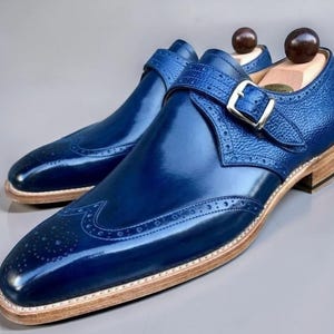 May include: A pair of blue leather dress shoes with a glossy finish. The shoes feature a monk strap with a silver buckle and decorative perforations. The soles are light brown with a wooden heel.