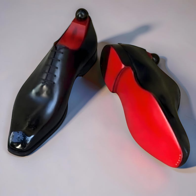 May include: A pair of black leather dress shoes with red soles. The shoes have a classic lace-up design and a glossy finish. The shoe trees are black with a round top.