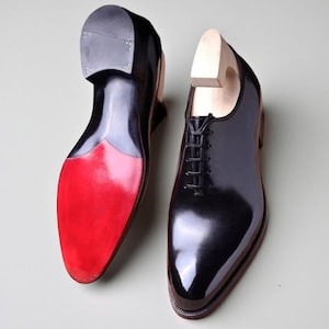 May include: A pair of black leather dress shoes with a high-gloss finish. The soles are red, and the shoes feature a traditional lace-up design. Wooden shoe trees are inserted inside the shoes.