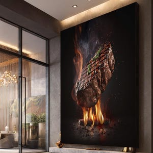 Op de afbeelding: Een groot, verticaal canvasdoek met een gegrilde steak met vlammen en rook tegen een zwarte achtergrond. De steak is gegarneerd met rozemarijn. De print wordt gepresenteerd in een modern interieur met glazen deuren.