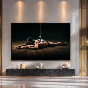 May include: A large flat-screen television displays a dark image of a person in black lingerie, lying on a wooden floor with a pole. The TV is mounted on a wall with gold accents, above a dark stone console.