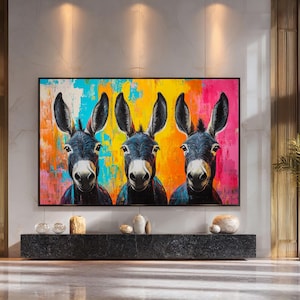 May include: A colourful framed print depicting three donkeys with black fur and large ears. The background is a vibrant mix of colours, including yellow, orange, turquoise, and pink. The artwork is displayed above a dark grey rectangular console.