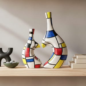 May include: Two abstract ceramic vases with a white base, featuring a colourful geometric design in red, yellow, blue, and black. The vases have a unique, curved shape and are displayed on a wooden shelf.