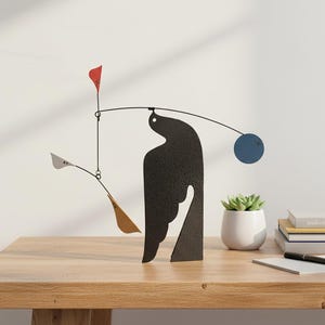 May include: A contemporary black metal mobile sculpture featuring a bird-like form, a red flag, a blue disc, and gold and silver shapes. The sculpture is on a wooden table with a small potted plant and books.