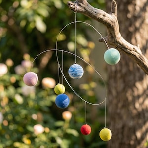 May include: A modern mobile with colorful felt spheres in shades of pink, blue, green, yellow, and red. The spheres are suspended from a silver wire frame, creating a whimsical and artistic display. The mobile is hanging from a tree branch.