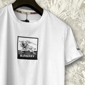 Puede incluir: Camiseta blanca con un gráfico en blanco y negro de un caballero a caballo dentro de un rectángulo negro. La palabra "BURBERRY" está impresa debajo. La camiseta tiene mangas cortas y cuello redondo.