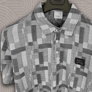 Puede incluir: Un polo de manga corta con un patrón geométrico en tonos grises y blancos. La camisa presenta un cuello, una tapeta abotonada y una pequeña etiqueta de marca. Un parche de marca es visible en la parte inferior derecha.