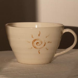 Puede incluir: Taza de cerámica color crema con un diseño de sol marrón. La taza tiene forma redondeada, un asa y una superficie lisa. El diseño del sol está pintado a mano y añade un toque decorativo.