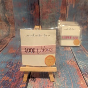 Könnte beinhalten: Ein kleiner Holzstaffelei zeigt eine Karte mit einem rosa Band mit der Aufschrift "GOOD Vibes". Die Karte befindet sich in einer transparenten Plastikhülle. Über dem Band steht "made with love". Im Hintergrund ist eine zweite Karte zu sehen.