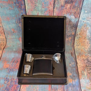 Könnte beinhalten: Ein schwarzes, lederbezogenes Flachmann-Set mit zwei Schnapsgläsern und einem Trichter, präsentiert in einer schwarzen Box mit weißer Naht. Das Set ist zum Tragen und Genießen von Spirituosen konzipiert.