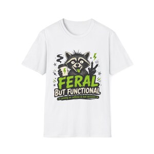 Könnte beinhalten: Weißes T-Shirt mit einer Grafik eines Waschbären, der eine Kaffeetasse hält und das Peace-Zeichen zeigt. Der Text "FERAL BUT FUNCTIONAL" ist grün, darunter steht "running on caffeine & bad decisions".