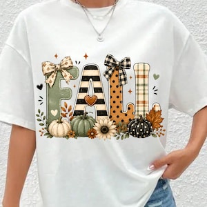 Op de afbeelding: Een wit t-shirt met het woord FALL in grote, decoratieve letters. De F is saliegroen met een strik, de A heeft zwarte strepen, de L is oranje met zwarte stippen en de tweede L heeft een ruitpatroon. Pompoenen en zonnebloemen sieren de onderkant.