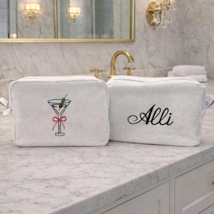 May include: Two white cosmetic bags with a textured weave. One bag features a martini glass embroidery with a pink bow, and the other has the name "Alli" embroidered in black script. The bags are displayed on a marble countertop.
