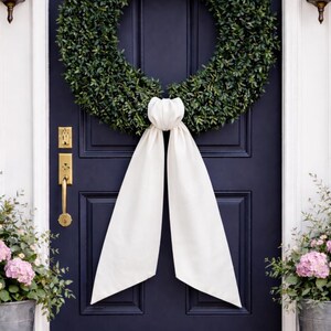May include: A lush green wreath hangs on a navy blue door, adorned with a large white fabric bow. The door features gold hardware and is flanked by potted plants with pink flowers, creating a welcoming entrance.