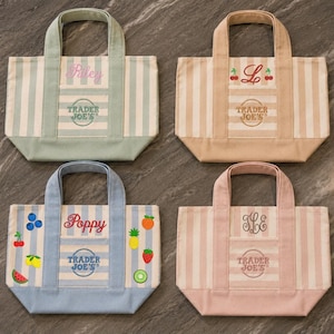 May include: Four small canvas tote bags with vertical stripes and solid colored handles and bases. Each bag features an embroidered Trader Joe's logo and personalized text or fruit graphics, including names like Riley and Poppy, a letter L, and a monogram.
