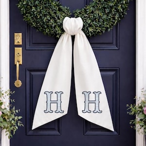 May include: A cream-coloured door sash with two embroidered "H" monograms in light blue and grey, hanging on a navy blue door. A green wreath is above the sash. The door has gold hardware.