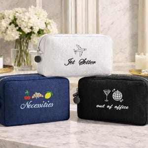 May include: Three rectangular zippered pouches with a textured fabric finish. The white pouch features an airplane icon and the text Jet Setter. The blue pouch displays a cherry, croissant, and lemon icon with the text Necessities. The black pouch shows a martini glass and disco ball icon with the text out of office.