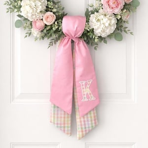 Op de afbeelding: Een bloemenkrans met roze rozen, witte hortensia's en groen. Een roze satijnen strik met een pastelkleurige geruite staart en een wit geborduurde letter "K" hangt aan de krans. De krans wordt op een witte deur getoond.