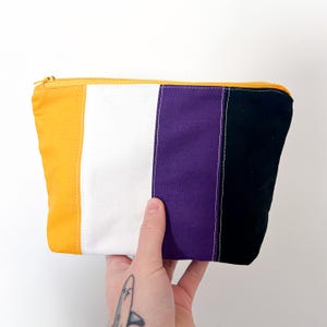 Könnte beinhalten: Ein rechteckiger Beutel mit Reißverschluss und Farbblock-Design. Der Beutel hat vertikale Streifen in Gelb, Weiß, Lila und Schwarz. Der Reißverschluss ist gelb. Der Beutel wird von einer Hand gehalten.