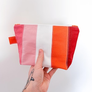 May include: A rectangular, striped pouch in shades of pink, white, orange, and red. The pouch has a small orange fabric loop on the side and a white zipper. The pouch is held by a hand with a tattoo.