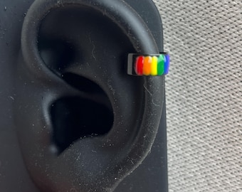 Könnte beinhalten: Ein schwarzer Ear Cuff mit einem leuchtenden, regenbogenfarbenen Emaille-Streifen. Das Schmuckstück ist auf einem schwarzen, ohrförmigen Ständer befestigt und zeigt eine glatte Oberfläche sowie ein kräftiges, mehrfarbiges Design am äußeren Rand.