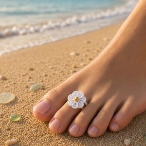 May include: A white crocheted flower toe ring featuring a small, smooth, metallic gold-colored bead at its center. The ring is constructed from fine white thread, forming a six-petaled floral shape that sits securely on the second toe of a foot.
