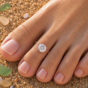 May include: A clear, transparent toe ring featuring a single, round, brilliant-cut white gemstone set in a silver-toned prong setting, worn on the second toe of a foot.