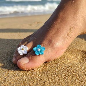 May include: Two crochet flower toe rings, one in white with a gold-toned center bead and one in bright blue with a white center bead. Each ring features a five-petaled floral design crafted from fine thread, worn on the toes of a foot.