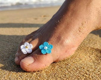 May include: Two crochet flower toe rings, one in white with a gold-toned center bead and one in bright blue with a white center bead. Each ring features a five-petaled floral design crafted from fine thread, worn on the toes of a foot.