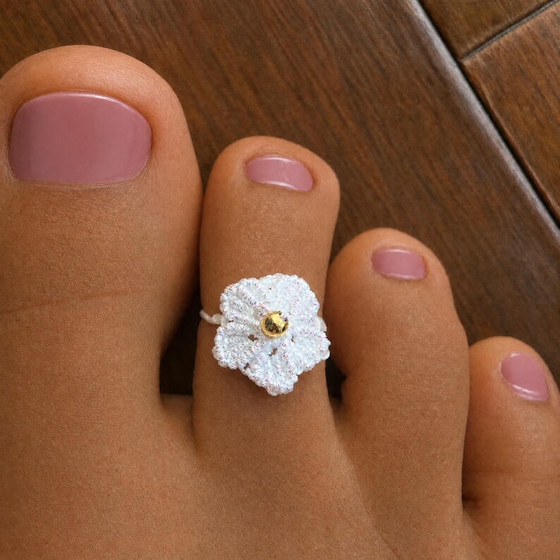 May include: A white crocheted flower toe ring featuring a small, metallic gold-colored bead at its center. The floral design is crafted from fine white thread and is positioned on the second toe, which has a light pink nail polish.