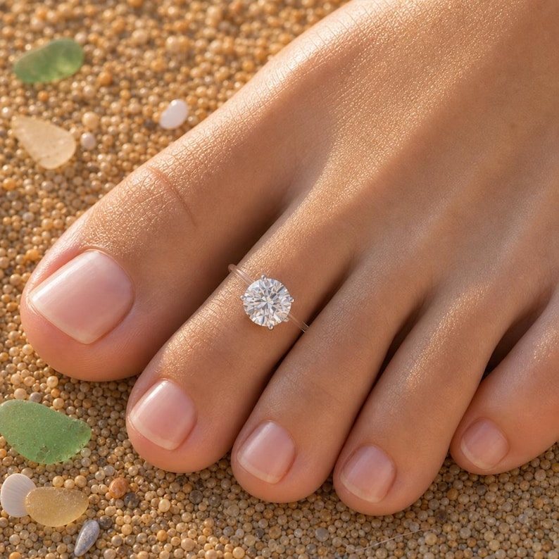 May include: A clear, transparent toe ring featuring a single, round, brilliant-cut white gemstone set in a silver-toned prong setting, worn on the second toe of a foot.