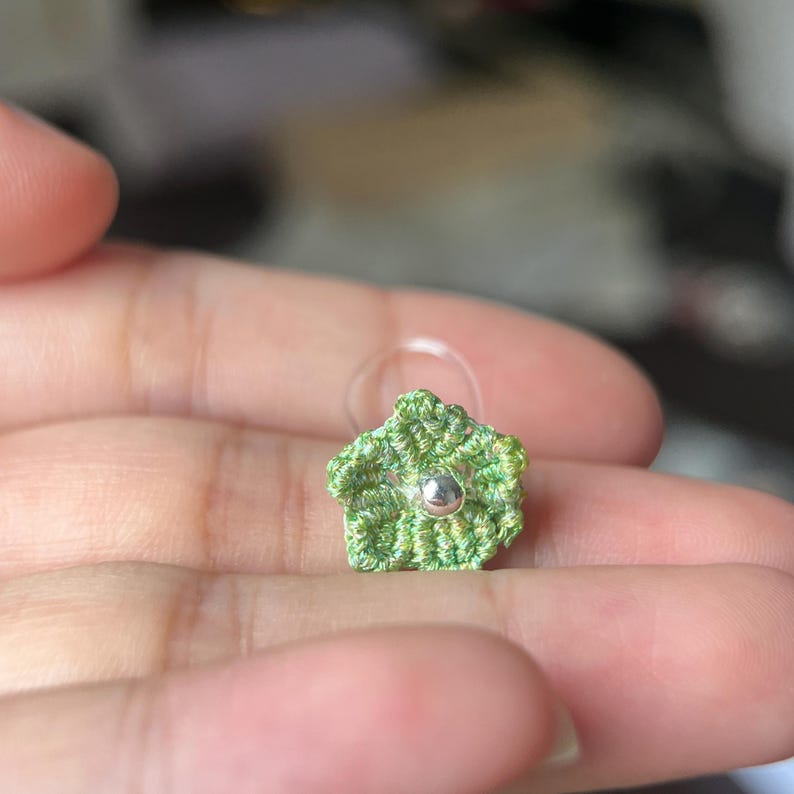 May include: A small, five-petaled flower stud earring crafted from light green thread with a metallic silver bead at the center. The floral design features a textured, crocheted appearance and includes a clear plastic backing for secure attachment.