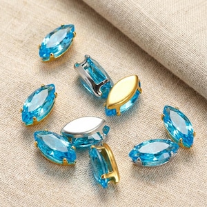 以下が含まれることがあります： 金と銀のセッティングが施された、マーキス型のライトブルーの宝石が、ニュートラルな生地の上に散らばっています。宝石はファセット加工されており、光を反射します。セッティングの色が異なり、コントラストを生み出しています。ジュエリー作りに適しています。