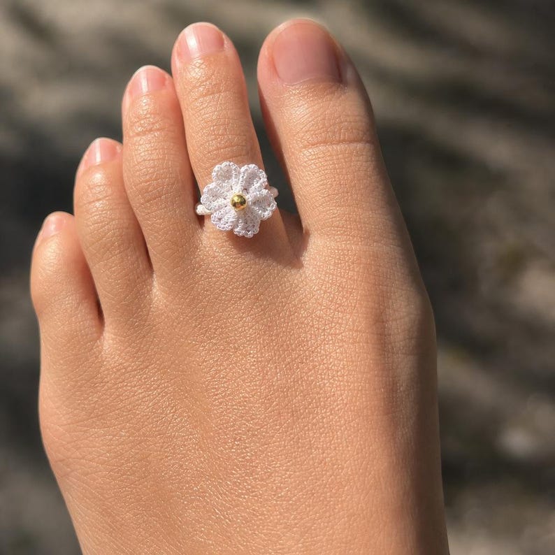 May include: A white crocheted flower toe ring featuring a small, smooth, metallic gold-colored bead at its center. The ring is constructed from fine white thread, forming a six-petaled floral design that encircles the second toe of a foot.