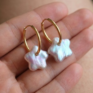 May include: A pair of gold hoop earrings with star-shaped, iridescent, freshwater pearls. The pearls have a soft, pearlescent sheen with hints of pink and blue. The earrings are displayed against a blurred background.