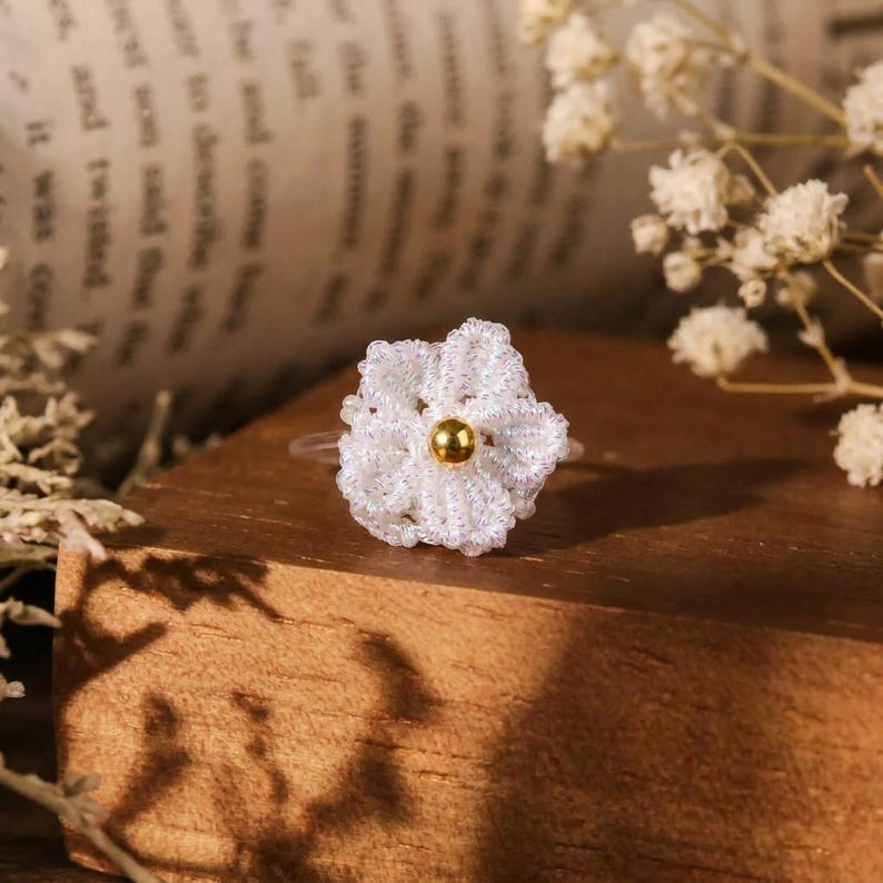 May include: A ring featuring a white crocheted flower motif with a small, smooth, gold-toned metallic bead at its center. The flower is constructed from fine, slightly iridescent thread, and the ring band is made of a thin, clear material.