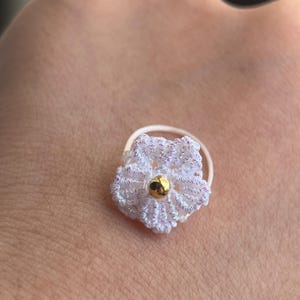 May include: A small, circular ring featuring a white crocheted flower with subtle iridescent highlights. The center of the flower is adorned with a single, smooth, polished gold-toned bead. The band of the ring is thin, white, and circular.