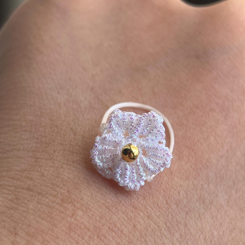 May include: A small, circular ring featuring a white crocheted flower with subtle iridescent highlights. The center of the flower is adorned with a single, smooth, polished gold-toned bead. The band of the ring is thin, white, and circular.