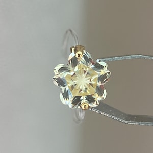 May include: A flower-shaped ring featuring a pale yellow, faceted gemstone set in a gold-toned metal prong setting. The ring band is thin and clear, providing a subtle appearance that highlights the central floral design and its reflective facets.