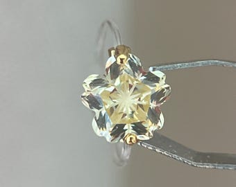 May include: A flower-shaped ring featuring a pale yellow, faceted gemstone set in a gold-toned metal prong setting. The ring band is thin and clear, providing a subtle appearance that highlights the central floral design and its reflective facets.