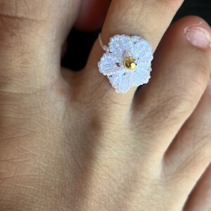 May include: A white crocheted flower toe ring featuring a small, shiny gold-toned bead at its center. The delicate, thread-based floral design has a subtle iridescent sheen and is worn around the second toe, showcasing intricate needlework.