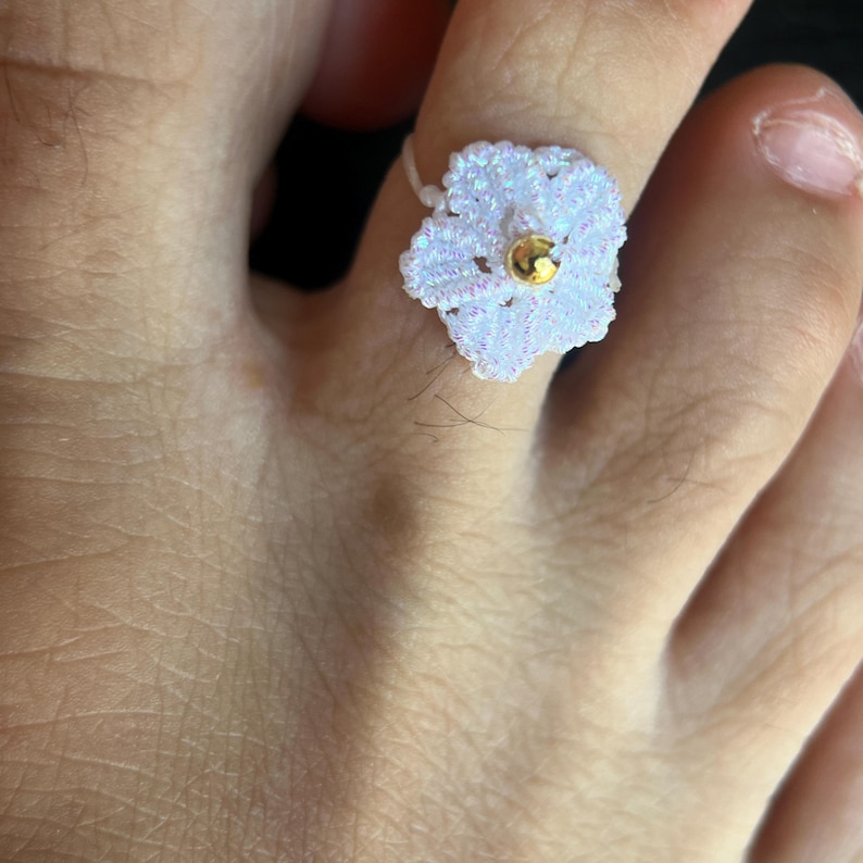 May include: A white crocheted flower toe ring featuring a small, shiny gold-toned bead at its center. The delicate, thread-based floral design has a subtle iridescent sheen and is worn around the second toe, showcasing intricate needlework.