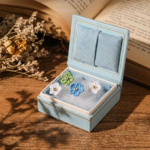 May include: A light blue velvet jewelry box containing four small crocheted flower pins. The pins are white, light green, and blue, each featuring a small metallic bead at the center. The box is open, revealing a white cushioned interior lining.