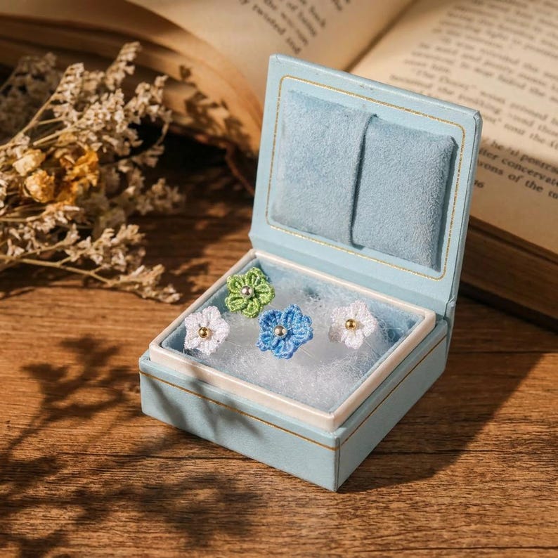May include: A light blue velvet jewelry box containing four small crocheted flower pins. The pins are white, light green, and blue, each featuring a small metallic bead at the center. The box is open, revealing a white cushioned interior lining.