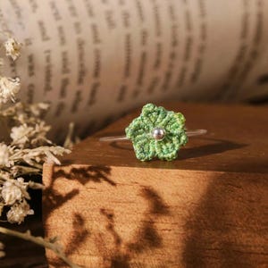 May include: A ring featuring a small, five-petaled flower crocheted from variegated green thread. The center of the flower is adorned with a single, smooth, polished silver-toned metal bead. The band of the ring is thin, clear, and circular.