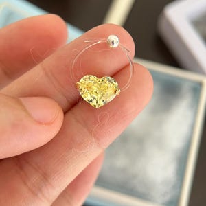 May include: A necklace featuring a single heart-shaped yellow gemstone set in gold-toned metal, suspended on a thin, transparent nylon cord. The cord is secured with a small, round, silver-toned bead clasp at the top, creating a floating effect.