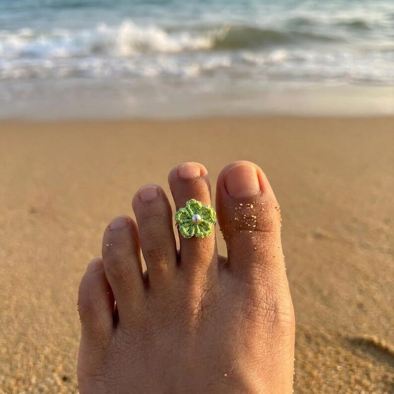 May include: A lime green crocheted flower toe ring featuring a small, centered silver-toned bead. The floral design is crafted from fine thread, creating a textured, petal-like appearance that sits securely around the toe.