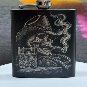 May include: A matte black metal flask featuring a laser-engraved illustration of a skull wearing a cowboy hat, smoking a cigar, and positioned next to a glass of whiskey with ice and a coiled rope. The flask has a silver-toned screw-top cap.