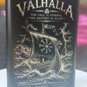 May include: A matte black metal flask featuring a laser-engraved Viking longship on a stormy sea. The text reads VALHALLA, THE CALL IS ETERNAL, THE DESTINY IS GLORY, and LIVE BRAVE, DIE HONORED, REACH VALHALLA, with a silver cap at the top.